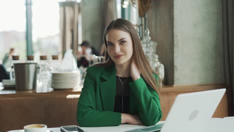 Una-Mujer-Encantadora-Con-Una-Chaqueta-Verde-Mira-La-Cámara-Y-Sonríe-Con-El-Telón-De-Fondo-De-Un-Hermoso-Restaurante