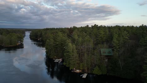 Aerial-view-of-a-calm-river-winding-through-dense-forest-with-cabins-along-the-banks-and-an-overcast-sky