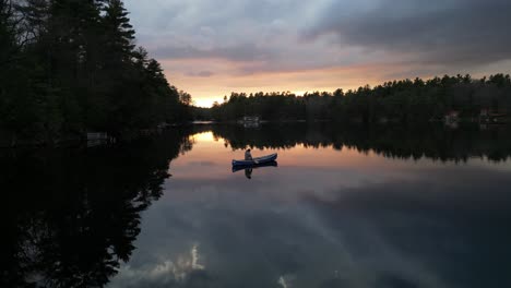 Eine-Person,-Die-Bei-Sonnenuntergang-Auf-Einem-Stillen-See-Kanu-Fährt,-Während-Sich-Die-Leuchtenden-Farben-Des-Himmels-Im-Wasser-Spiegeln,-Umgeben-Von-Dichten-Bäumen