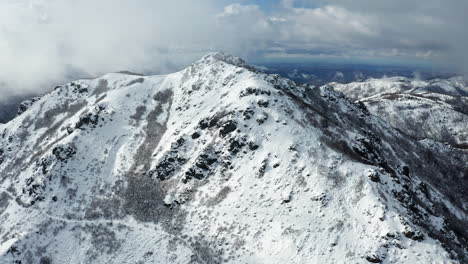 Snow-capped-mountain-peaks-under-a-cloudy-sky,-aerial-view-captures-the-rugged-terrain-and-vast-wilderness