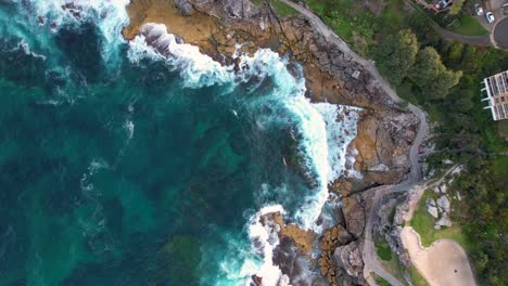 A-birds-eye-drone-view-looking-down-as-the-blue-ocean-waves-crash-against-the-rocks-and-the-drone-is-rising-to-reveal-more-of-the-Australian-landscape-around