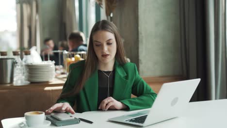 Una-Joven-Profesional-Caucásica-Está-Trabajando-En-Su-Computadora-Portátil-En-Un-Café,-Pero-Está-Constantemente-Molesta-Y-Distraída-Por-Su-Teléfono-Inteligente.