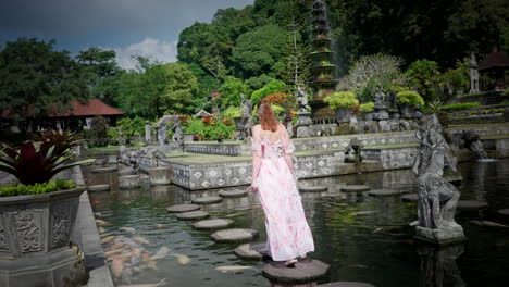 Female-tourist-in-summer-dress-walk-over-Tirta-Gangga-stepping-stones,-Bali