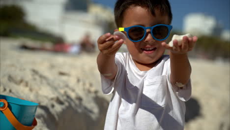 Cámara-Lenta-De-Un-Niño-Latino-Mexicano-Con-Gafas-De-Sol-Y-Una-Camiseta-Blanca-Mirando-La-Cámara-Mostrando-Algunas-Conchas-Marinas-Que-Encontró-En-La-Playa-De-Cancún