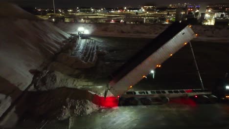 Nighttime-dump-truck-unloading-sand-at-a-Montreal-construction-site,-city-lights-in-background,-aerial-shot