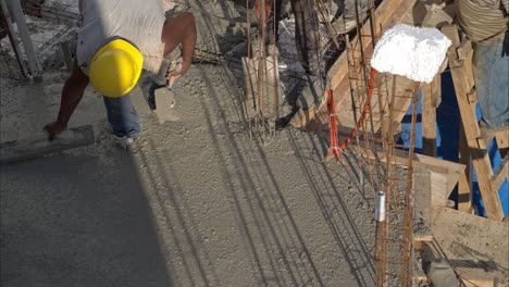Tilt-down-shot-of-an-mexican-latin-construction-worker-with-a-yellow-hardhat-flattening-fresh-concrete-using-a-wooden-trowel-on-a-sunny-afternoon-in-slow-motion