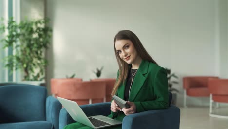 Una-Mujer-Encantadora-Con-Una-Computadora-Portátil-Y-Una-Computadora-Portátil-Brilla-En-El-Vestíbulo-De-Un-Centro-De-Negocios,-Sonriendo-Y-Mirando-A-La-Cámara