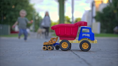 Unrecognizable-young-boy-running-from-his-mother-towards-the-camera-to-play-with-some-toy-trucks-placed-on-the-street
