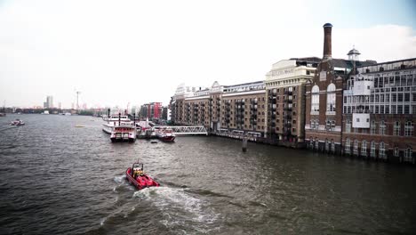 Un-Barco-Rojo-Vibrante-Navega-Por-Un-Río-Frente-A-Edificios-Históricos-En-Londres,-Mostrando-Un-Animado-Paisaje-Urbano-Junto-Al-Agua.