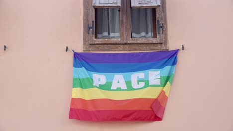 An-Italian-Peace-Flag-in-rainbow-colors-with-the-Italian-word-pace-on-it-hangs-beneath-a-window