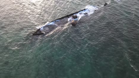 Drone-aerial-flying-low-over-a-shipwreck-in-the-ocean-with-waves-crashing-into-it