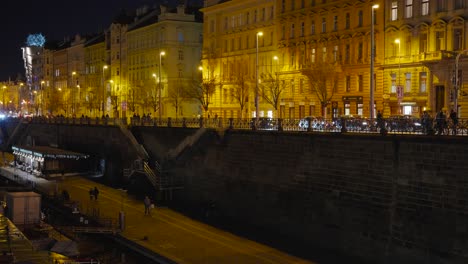 Pedestrian-sidewalk-near-river-embankment-and-buildings-illuminated,-Prague