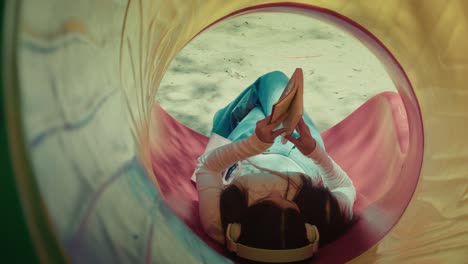 young-woman-listening-to-music-with-headphone-while-reading-book-novel-laid-on-park-slide