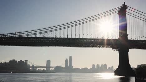New-York-City-Manhattan-Bridge-Von-Unten-Mit-Dem-East-River-Und-Der-Stadt-Im-Hintergrund-Mit-Sonnenaufgang,-Der-Durch-Die-Brücke-Scheint