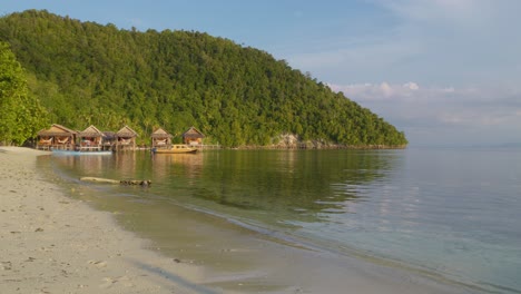 Tranquil-beach-on-Kri-Island-in-the-Raja-Ampat-Archipelago,-Indonesia,-featuring-wooden-huts-along-the-shore-against-a-lush-green-backdrop