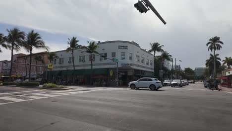 Busy-street-intersection-in-Miami-Beach-with-palm-trees,-shops,-and-pedestrians-on-a-sunny-day