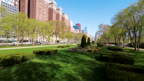 Luftaufnahmen-Einer-FPV-Drohne-Von-Einem-Ruhigen-Stadtpark-Mit-Gepflegten-Rasenflächen,-Blühenden-Blumen-Und-Gebäuden-Der-Stadt-Chicago-Unter-Einem-Klaren-Blauen-Himmel