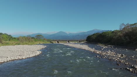 Dron-Cruzando-Un-Río-Con-Impresionantes-Aguas-Transparentes,-Dejando-Al-Descubierto-Un-Hermoso-Puente-Y-Las-Imponentes-Montañas-De-Los-Andes-Al-Fondo