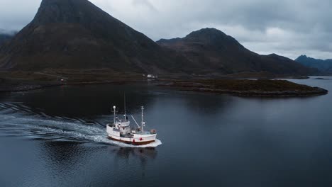 Aerial-view-orbiting-around-a-fisher-boat-returning-to-port-under-the-Flakstadbruene-bridge,-near-Fredvang,-Lofoten-Islands,-Norway