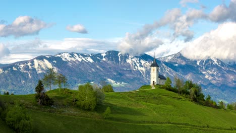 Malerische-Szene-Einer-Einsamen-Kirche-Auf-Einem-Grünen-Hügel,-Eingerahmt-Von-Der-Erhabenheit-Der-Schneebedeckten-Berge-Und-Einem-Himmel,-Der-An-Sonnigen-Tagen-Von-Schnell-Ziehenden-Wolken-Durchzogen-Ist