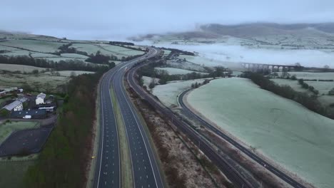 Flying-over-bridge-on-quiet-M6-motorway-towards-mist-covered-hills-at-dawn-in-winter-with-stone-viaduct-in-distance
