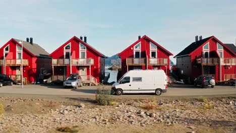 Lateral-tracking-shot-of-a-white-camper-van-in-front-of-gorgeous-red-rorbu-houses,-revealing-the-amazing-mountain-backdrop-of-Henningsvær,-Lofoten-Islands,-Norway