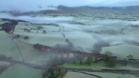 Derelict-stone-viaduct-surrounded-by-rolling-green-countryside-hills-at-dawn-in-winter-with-mist-and-low-cloud-cover
