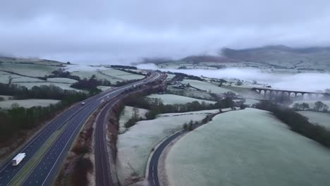 Light-traffic-on-M6-motorway-with-low-cloud-cover-on-hills-on-frosty-winter-morning-and-stone-viaduct-backed-by-mist-and-fog