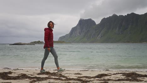 SLOW-MOTION,-a-young-woman-in-a-red-jacket-walks-on-a-beautiful-beach-in-front-of-the-crashing-waves-and-mountains-in-the-Lofoten-Islands,-Norway