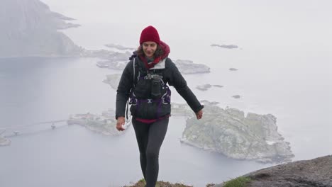 Una-Mujer-Atlética-Llegando-A-La-Cima-De-La-Caminata-Reinebringen-Con-Vistas-De-Hamnøy-Al-Fondo,-Reine,-Islas-Lofoten,-Noruega