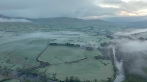 Flying-over-meandering-river-towards-mist-shrouded-patchwork-fields-at-dawn-in-winter