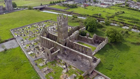 Claregalway-Abbey-ruins-grounds-with-empty-stone-arches,-drone-ascends-tilting-down