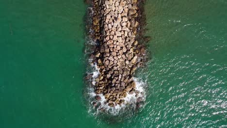 Rocky-pier-extending-into-turquoise-waters-in-sitges,-spain,-aerial-view