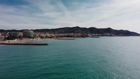 Sitges-coastline-with-city-buildings-and-mountains-in-the-background,-aerial-view