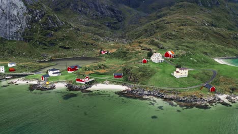 Aerial-view,-orbit-tilt-up,-a-few-red-rorbu-houses-in-a-tiny-coastline-fishing-village-at-the-foot-of-majestic-mountains-in-the-Lofoten-Islands,-Norway