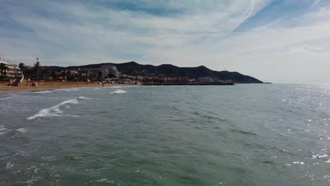 Vista-Panorámica-De-La-Costa-De-Sitges-Con-Olas-Suaves,-Un-Mar-En-Calma-Y-Montañas-Al-Fondo.