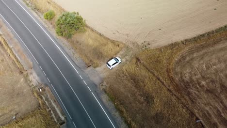 A-white-car-parked-next-to-a-road-in-a-dry-field,-aerial-view