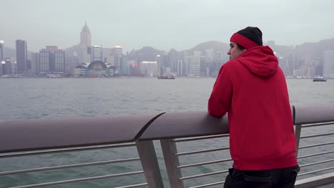 Ein-Junger-Mann-Steht-Auf-Einer-öffentlichen-Promenade-Und-Genießt-Die-Wunderschöne-Aussicht-Auf-Hongkong-Mit-Gebäuden-Und-Wolkenkratzern,-China-2020