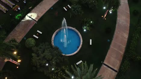 Toma-Aérea-De-Una-Pequeña-Fuente-De-Piscina-En-Un-Parque-Verdoso-Con-Luces-En-El-Césped