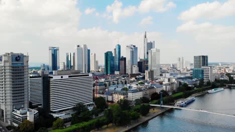 Aerial,-descending,-drone-shot-above-main-river,-towards-downtown-Frankfurt,-partly-sunny-day,-in-Germany---Quiet-due-to-Covid-19