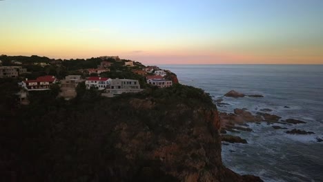 Volando-Sobre-Villas-Sentadas-En-El-Borde-Del-Acantilado-De-Knysna-Heads,-Hora-Dorada,-Sudáfrica,-Movimiento-Aéreo-Hacia-Adelante