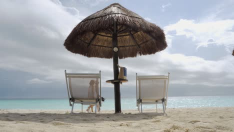 Strandliegen-Und-Sonnenschirm-Am-Meer-Mit-Einem-Paar,-Das-Am-Strand-Spazieren-Geht