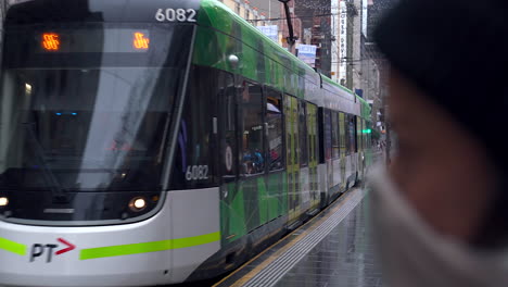 Melbourne-trams-pass-waiting-commuter-wearing-a-face-mask,-in-the-wet-city-center
