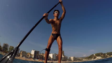 Un-Joven-En-Traje-De-Baño,-Remando-De-Pie-Con-La-Playa-De-La-Ciudad-Al-Fondo