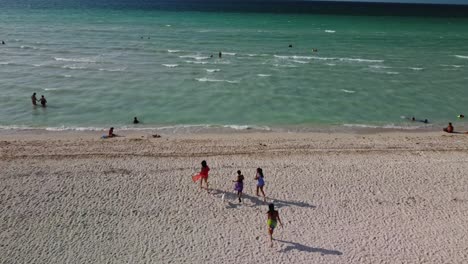 En-La-Vista-Superior-Tomada-Por-Un-Dron-Se-Puede-Observar-Un-Grupo-De-Amigos-Corriendo-En-La-Playa-De-Arena,-Turistas-Nadando-En-El-Agua-Clara-Y-Mareas-Espumosas.