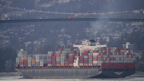 A-Cargo-Ship-Fully-Loaded-With-Container-Vans-Sailing-At-Brockton-Point,-Stanley-Park-In-Vancouver,-British-Columbia-With-Vehicles-Travelling-On-Lions-Gate-Bridge-In-The-Background---Medium-Shot