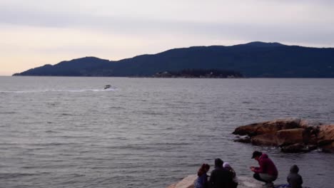 People-gather-at-coastline-sunset-with-speed-boat-passing,-long-shot