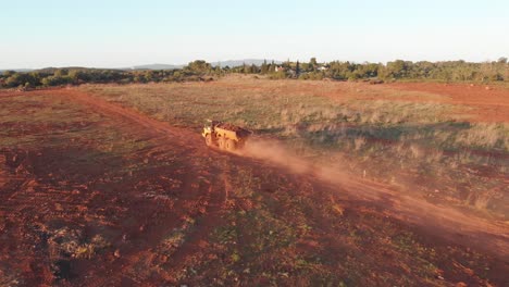 Vista-Aérea-De-Un-Camión-Volquete-Todoterreno-Con-Un-Remolque-Lleno-Conduciendo-Por-Un-Camino-De-Tierra-En-Un-Campo-Abierto-Durante-La-Puesta-De-Sol