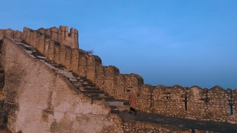 Vuelo-De-Drones-En-El-Fuerte-Ranikot-En-Sindh,-Pakistán---Hombre-Caminando-Sobre-La-Histórica-Pared-De-Ruinas