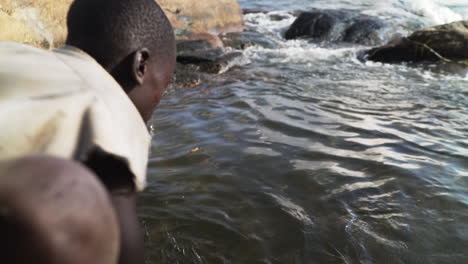 Un-Niño-Ugandés-Camina-Hasta-La-Orilla-Del-Río-Nilo-Y-Se-Lava-La-Cara-Con-Agua,-Uganda,-África